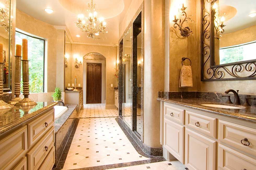 A luxurious, well-lit bathroom with beige cabinets, a chandelier, tiled flooring, and an arched doorway.