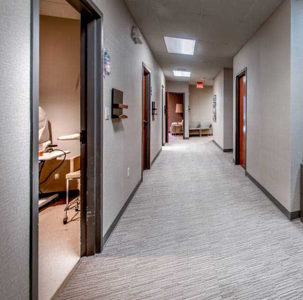 A hallway in a neutral-toned office with carpeted floors, multiple doorways, and an open room showing medical equipment.