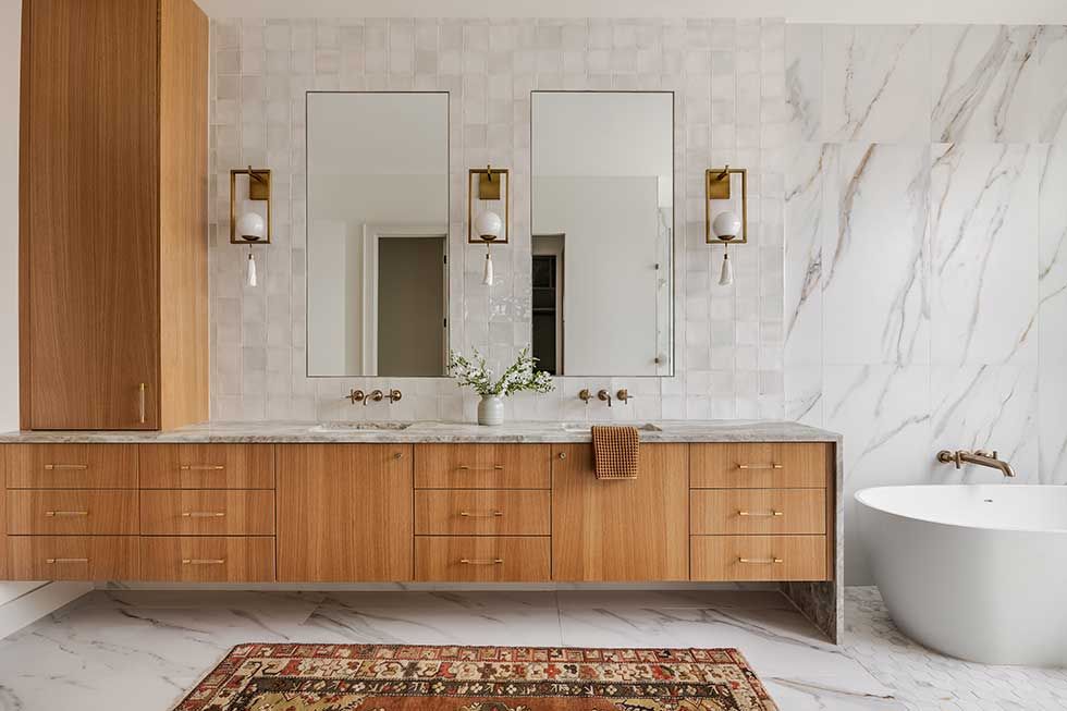 A double vanity bathroom with warm wood cabinets, white marble floors and walls, mirrors, and a freestanding bathtub.