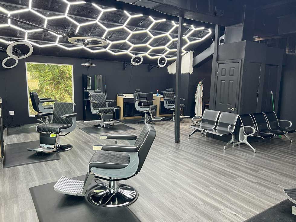 A modern barbershop with black walls, grey wood flooring, multiple barber chairs, and a hexagonal LED ceiling light.
