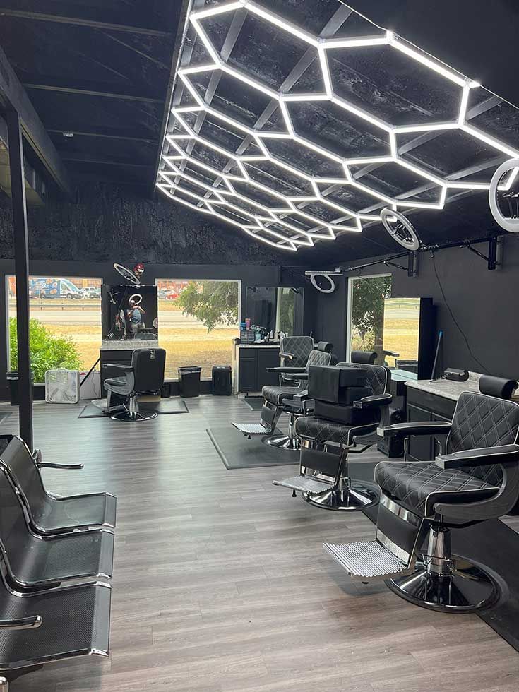 A modern barbershop with black walls, wood floors, leather chairs, and a geometric honeycomb LED ceiling light.