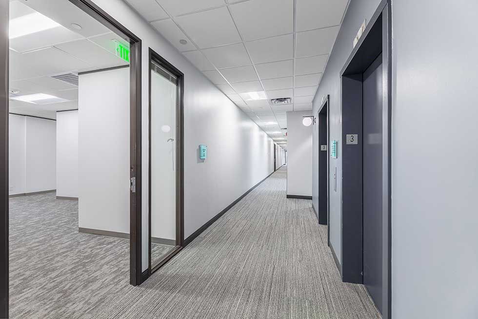 A long office hallway with light gray walls, carpeted floors, and a series of doors leading into rooms.
