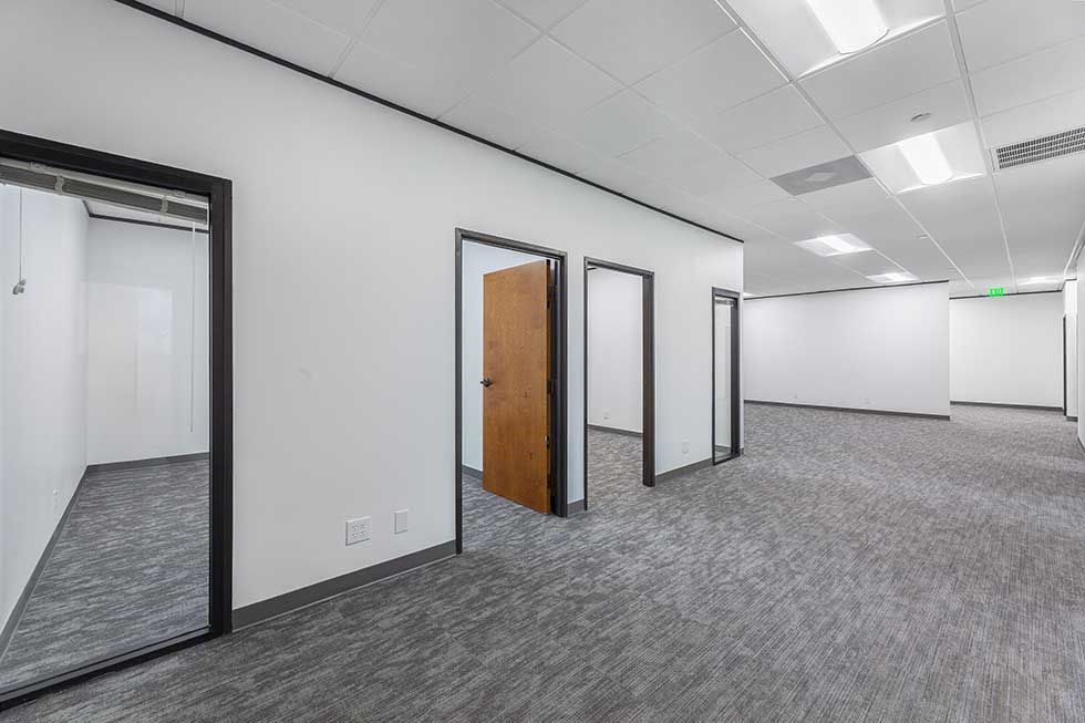 A bright, empty office hallway with grey carpet, white walls, and several doorways, including one with a wooden door.