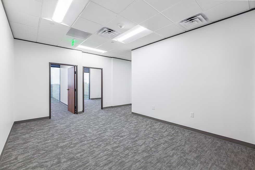 An empty office space with white walls, grey carpet, a drop ceiling with lights, and two open doorways.