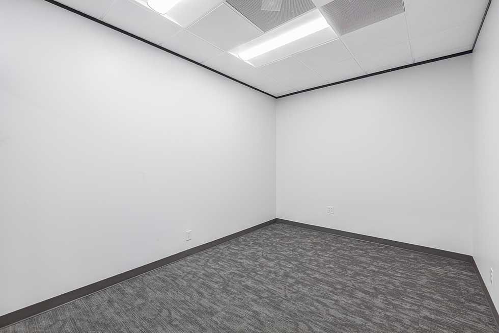 An empty office with white walls, gray carpet, and recessed ceiling lighting.