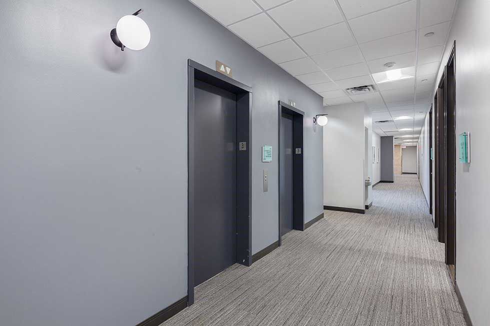 A hallway with gray walls, two dark elevator doors, a patterned carpet floor, and modern globe wall sconces.