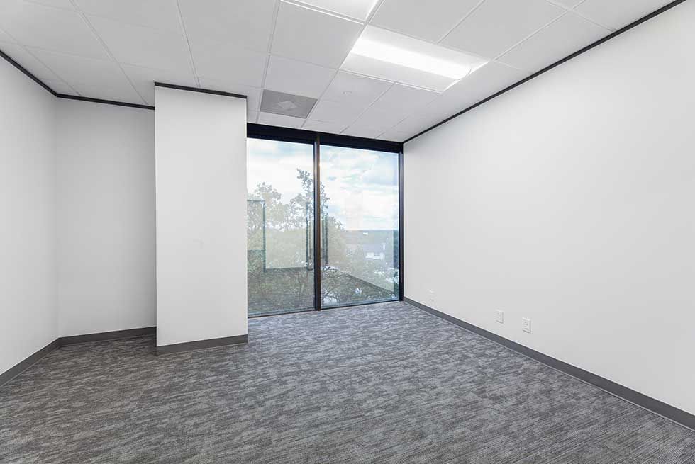 An empty office with white walls, gray patterned carpet, and a large window looking out at trees.