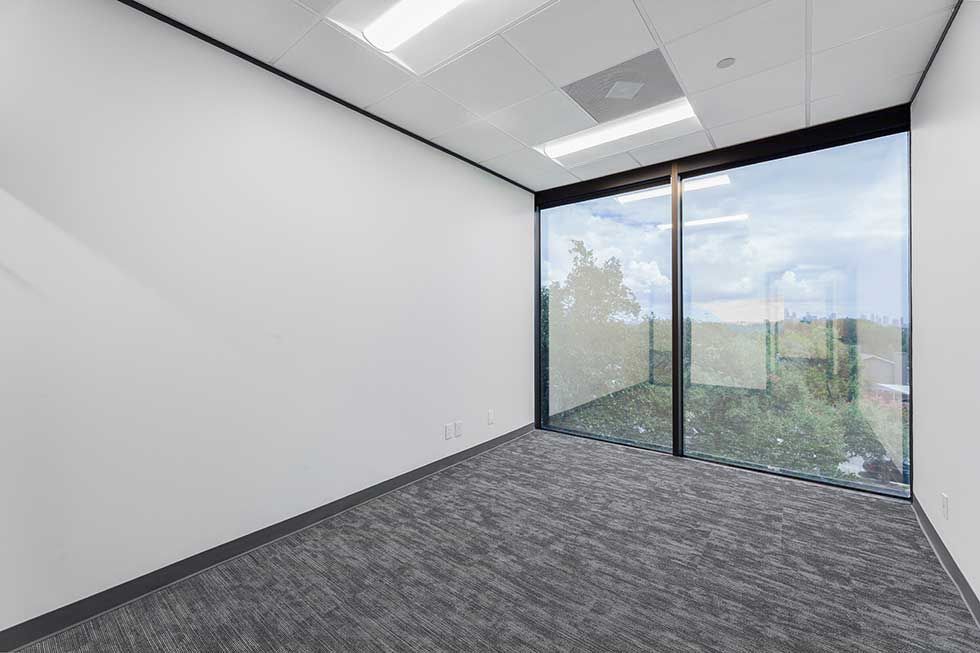 Empty office room with white walls, gray carpet, and floor-to-ceiling windows showing a view of green trees.