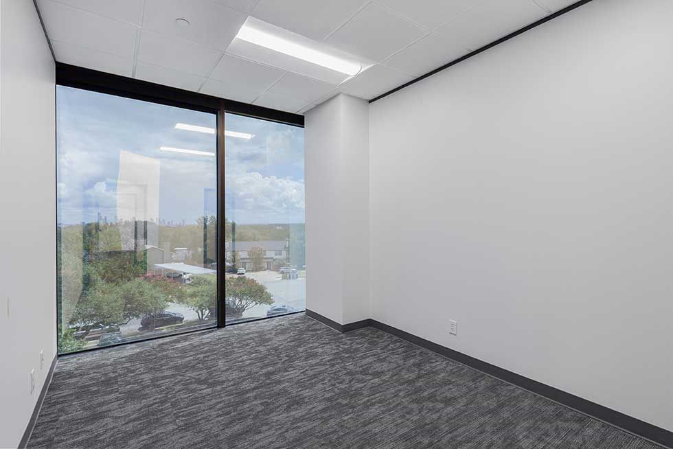 An empty, modern office space with white walls, grey patterned carpet, and a large window overlooking a tree-lined area.
