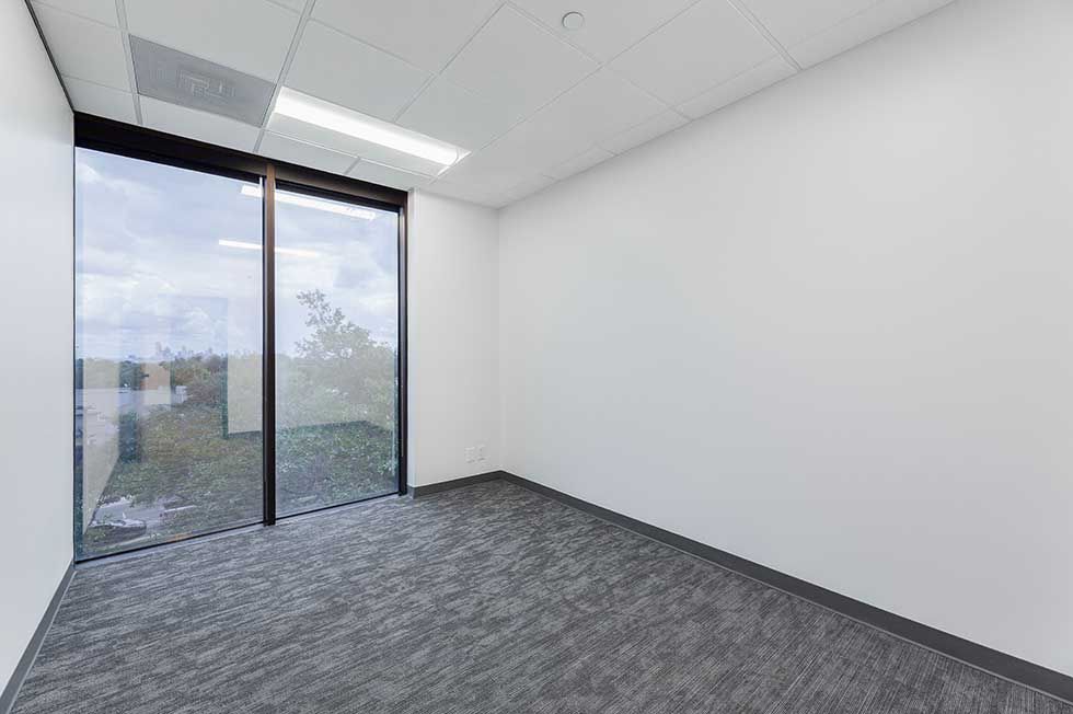 An empty office with white walls, gray carpet, a drop ceiling, and a large window looking out onto a grassy area.