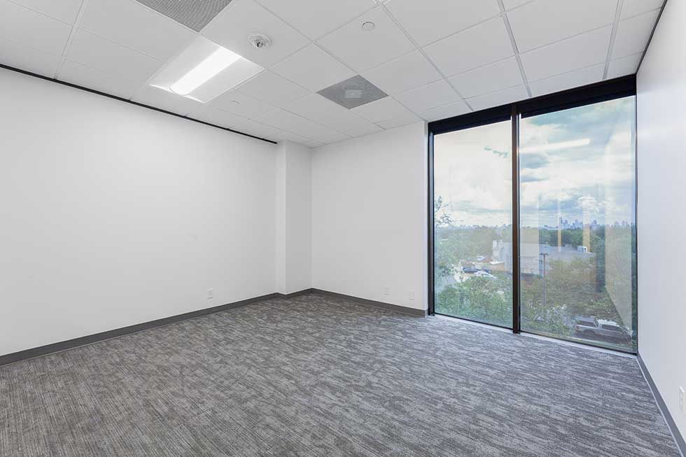 Empty office with white walls, gray carpet, a drop ceiling, and a floor-to-ceiling window overlooking trees and buildings.