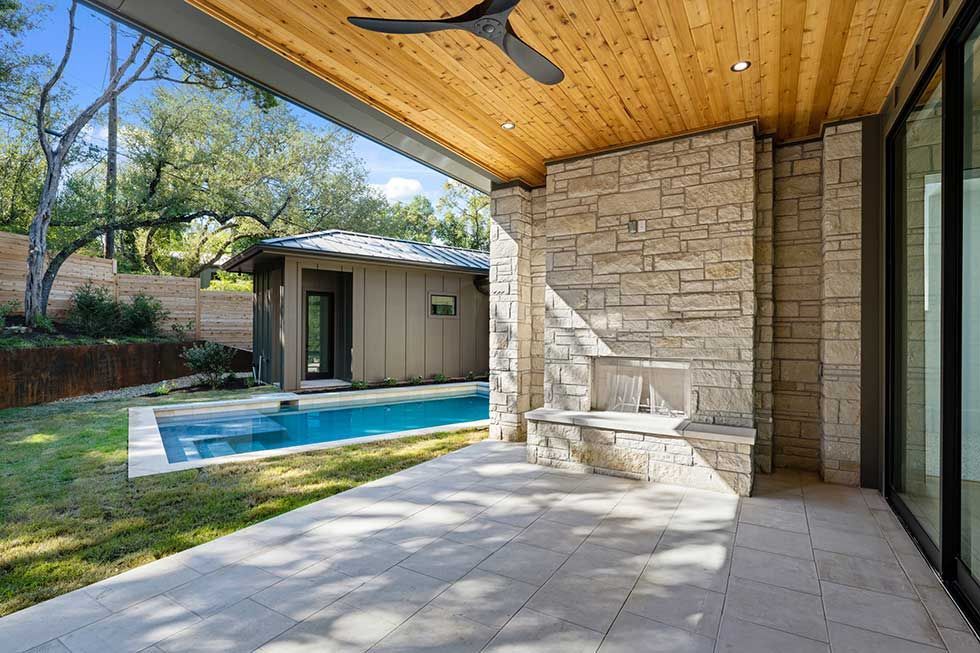 A stone fireplace on a shaded modern patio overlooking a backyard with a rectangular swimming pool and a small guest house.