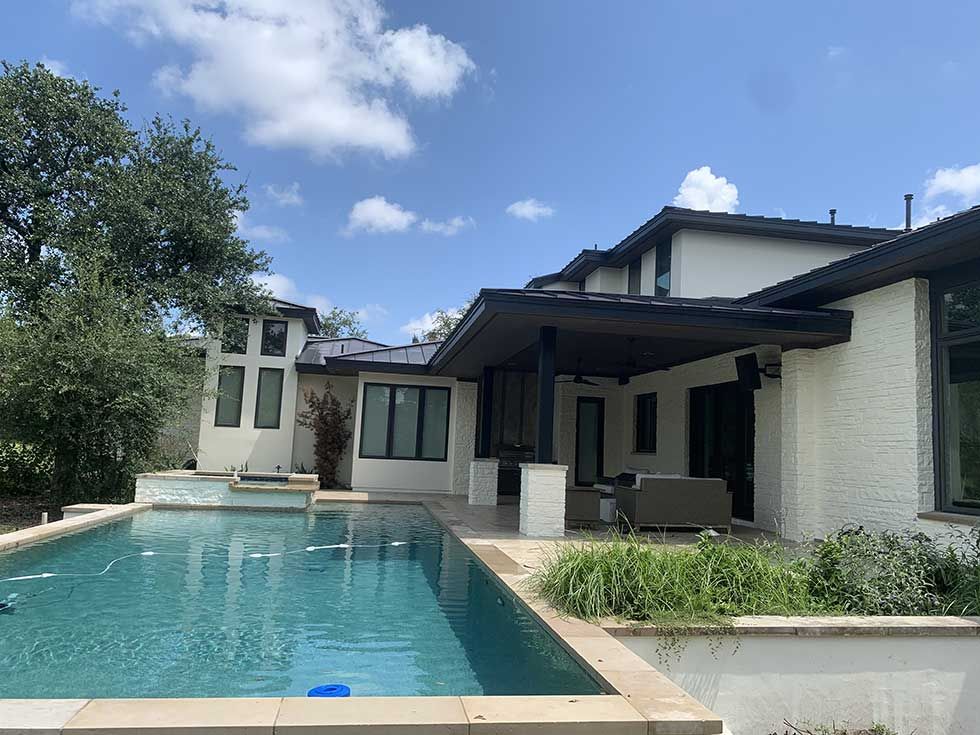 A modern white stucco house with a dark roof and a covered patio overlooking a swimming pool under a blue sky.