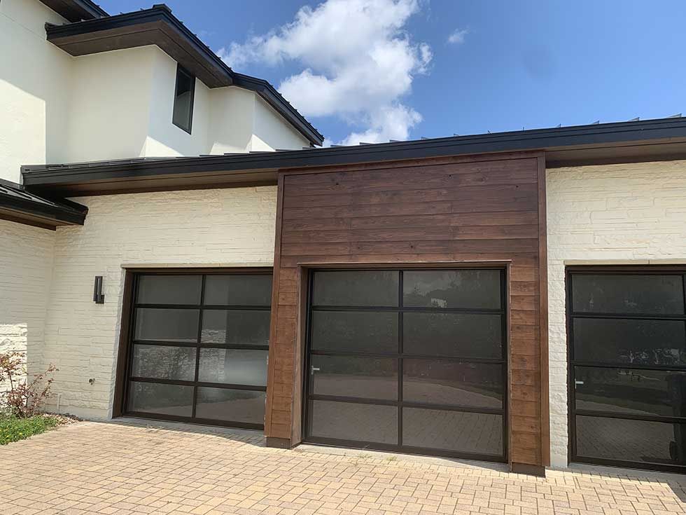Modern home exterior featuring a stone-clad wall with three glass-paneled garage doors beneath dark horizontal wood siding.