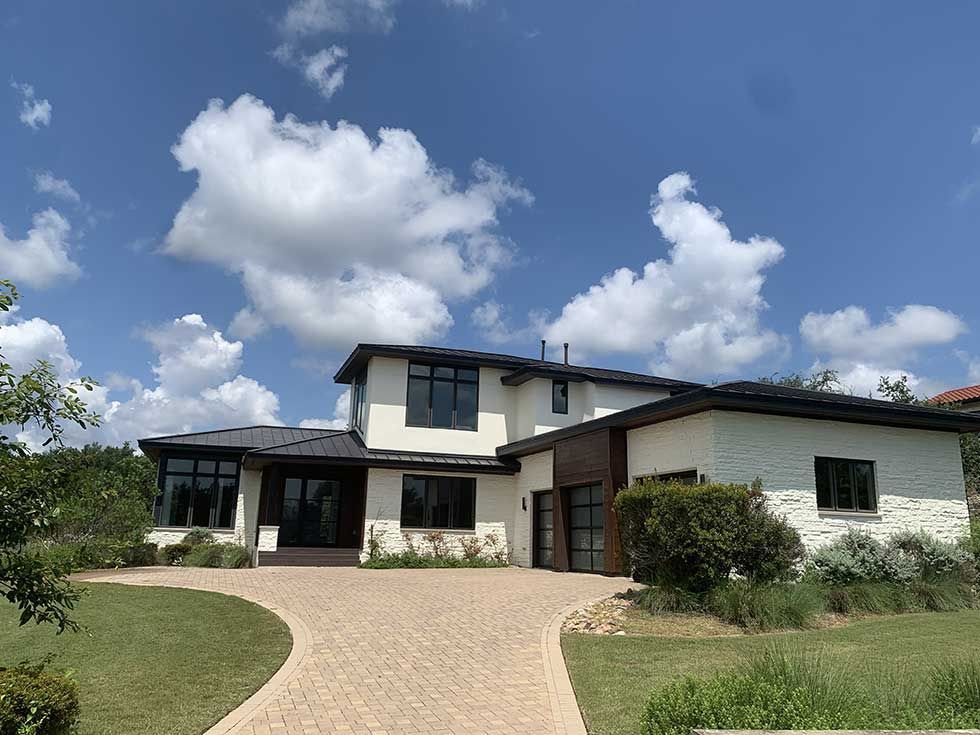 A modern two-story home with white stucco walls, dark wood accents, and a paved circular driveway under a blue sky.