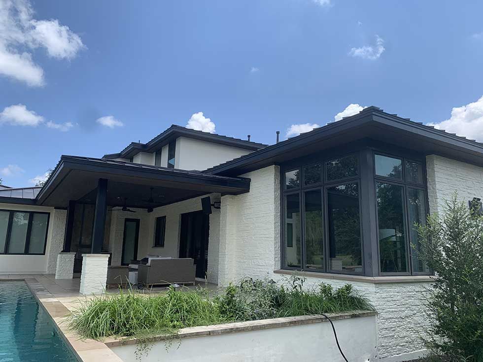 Modern white stone home with a dark roof, covered patio, and large windows overlooking a swimming pool on a sunny day.