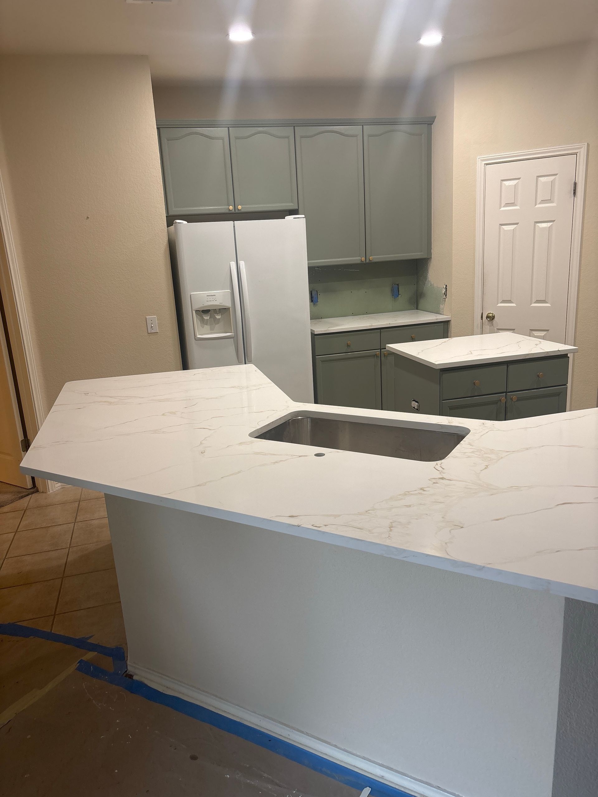 A kitchen island with a white marble countertop and sink, featuring grey cabinets and a white refrigerator in the background.