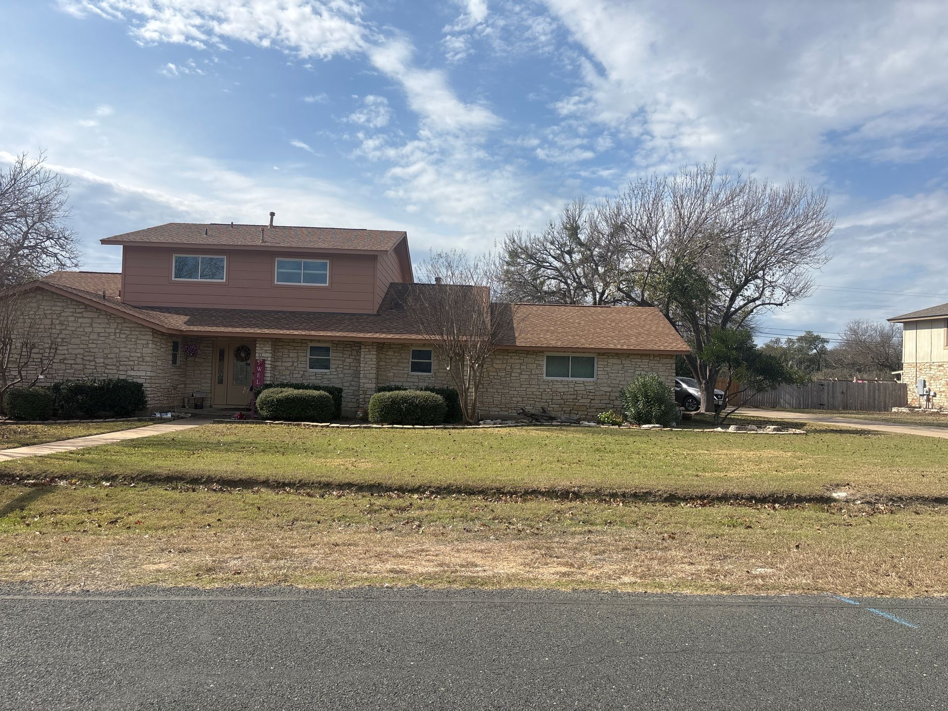 A two-story tan brick house with a brown shingled roof, brown upper siding, and a manicured lawn under a cloudy sky.