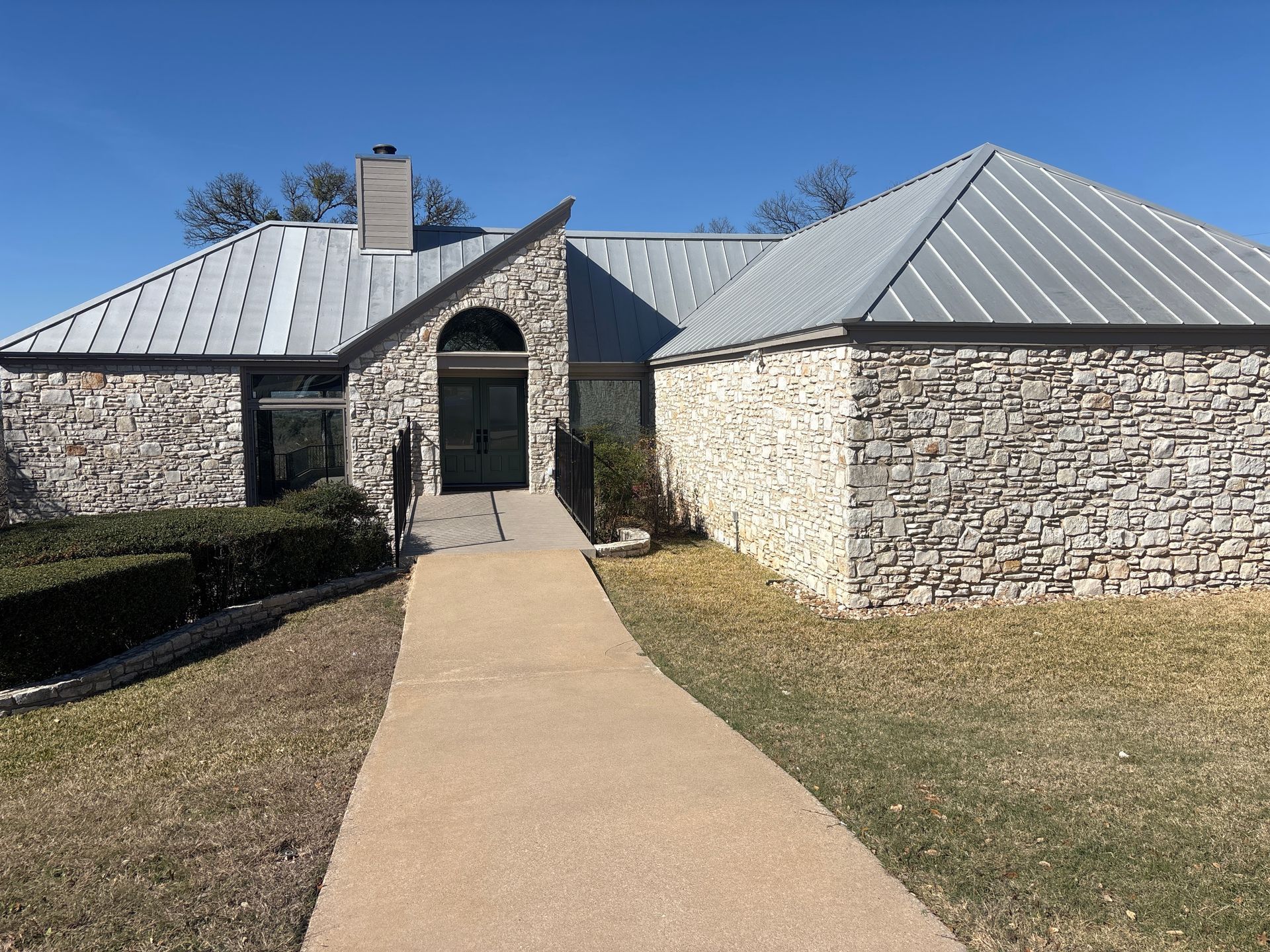 A stone house with a metal roof and a long concrete walkway leading to the front door under a clear blue sky.