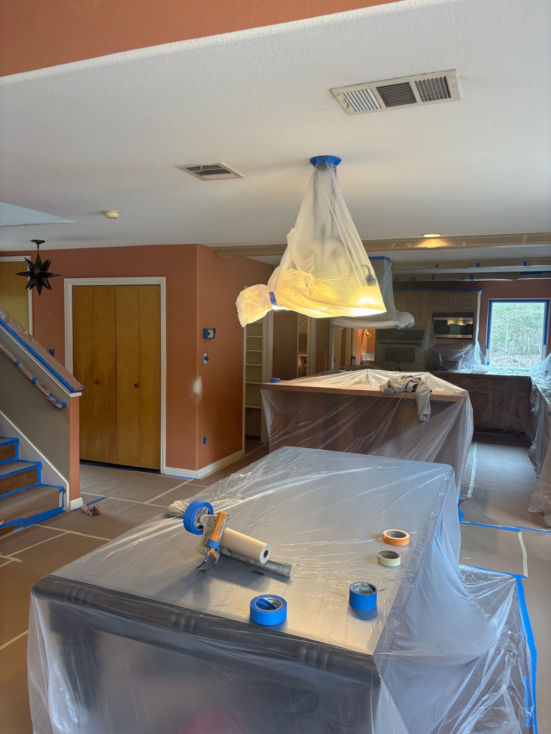 A kitchen interior covered in plastic sheets and painter's tape for painting preparation, with a chandelier draped in film.