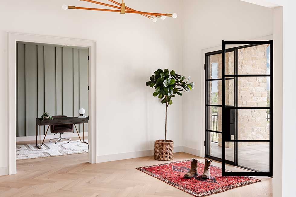 A bright foyer with herringbone wood floors, a potted fiddle leaf fig, an open office, and a black-framed glass door.