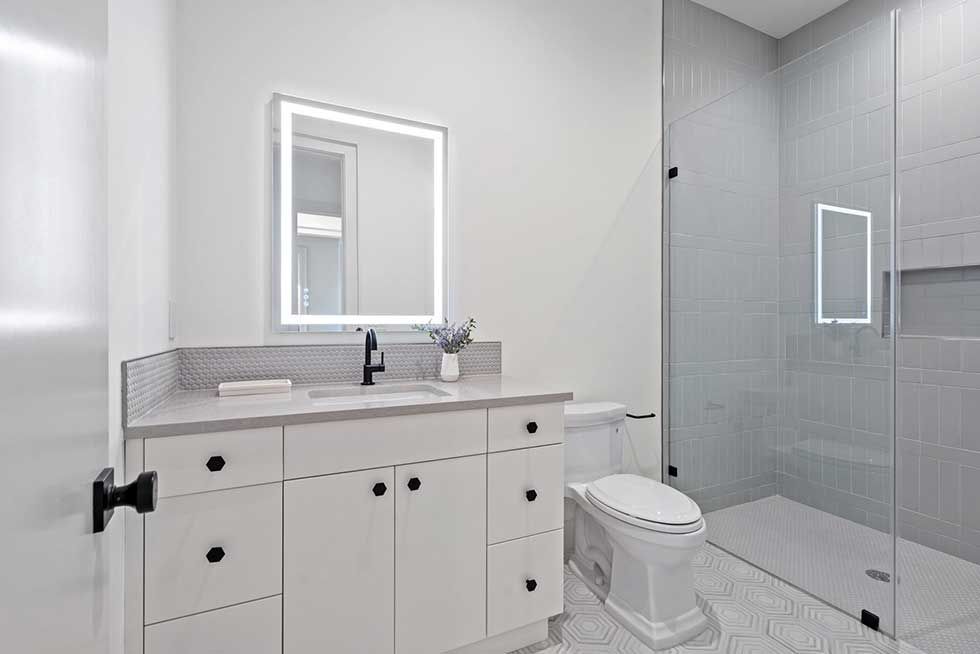 A modern bathroom with a white vanity, a backlit mirror, and a glass-enclosed shower with gray tiled walls.