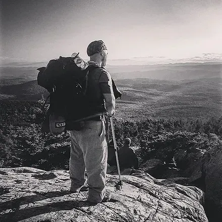 Hiker with backpack and walking stick atop a mountain, looking out over a valley on a sunny day.