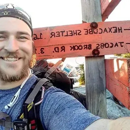 Man smiles near a wooden sign pointing to Hexacube Shelter and Jacobs Brook Rd. He wears a backpack and a hat.
