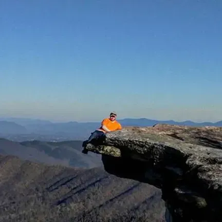 Man reclining on a rock ledge, with mountain range in background. Blue sky, sunny day.
