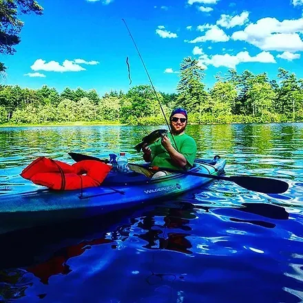 Man fishing from a blue kayak on a lake, holding a fish. Sunny day, forest background.