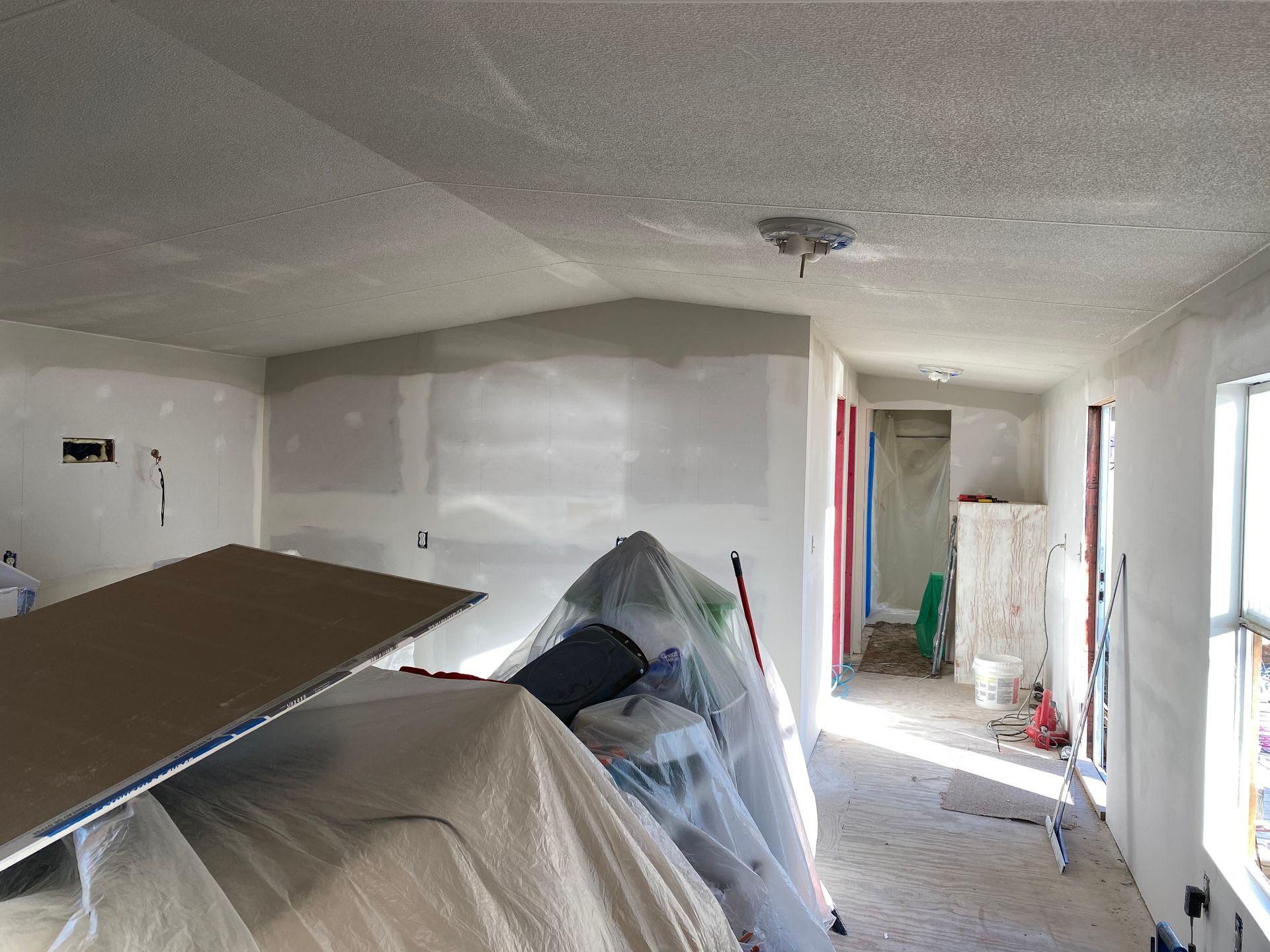 Interior of a room under construction, with drywall and covered materials; hallway visible in the background.