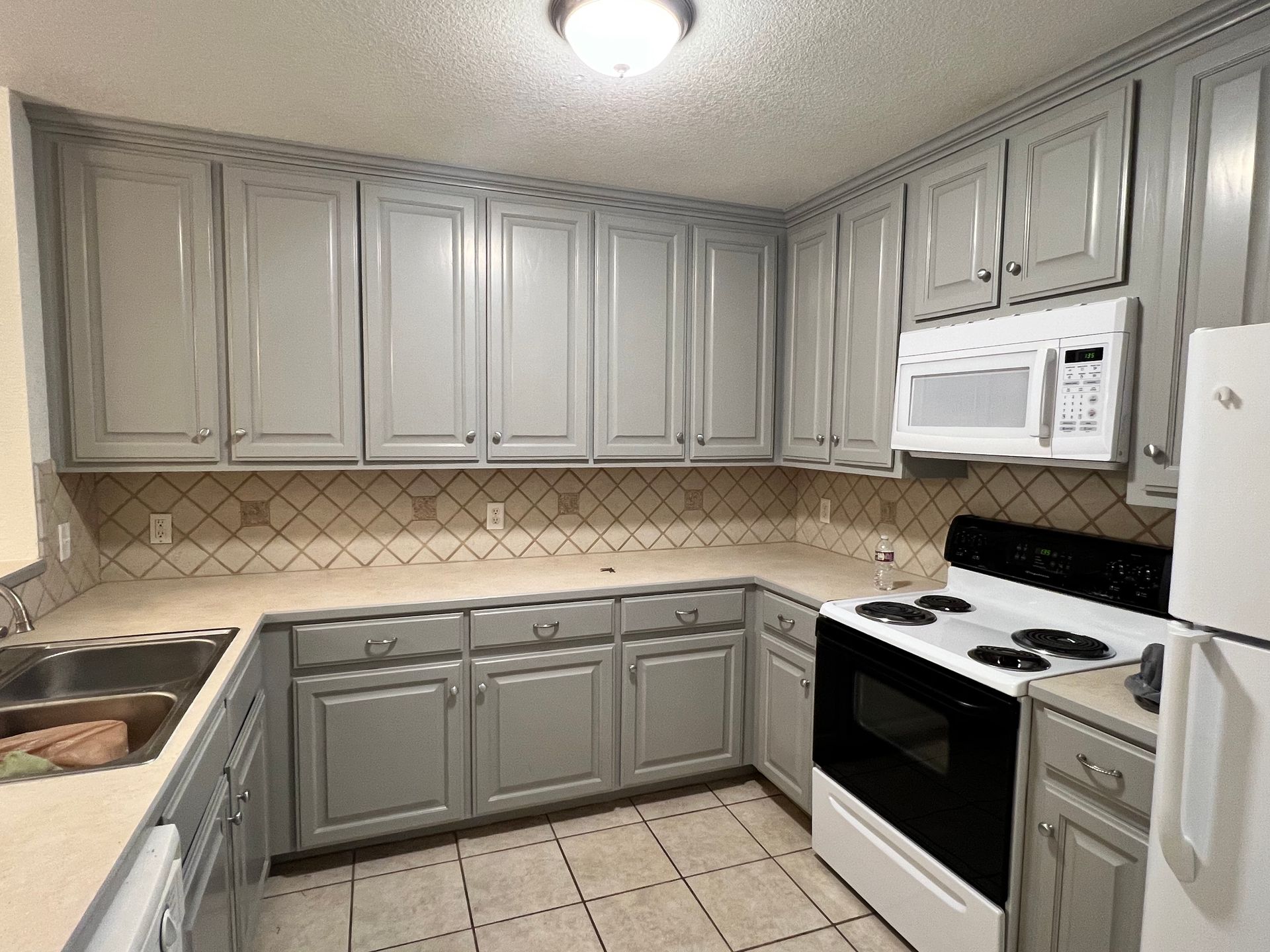 A kitchen with gray cabinets, a white microwave, stove, and refrigerator. Tiled backsplash and countertops.
