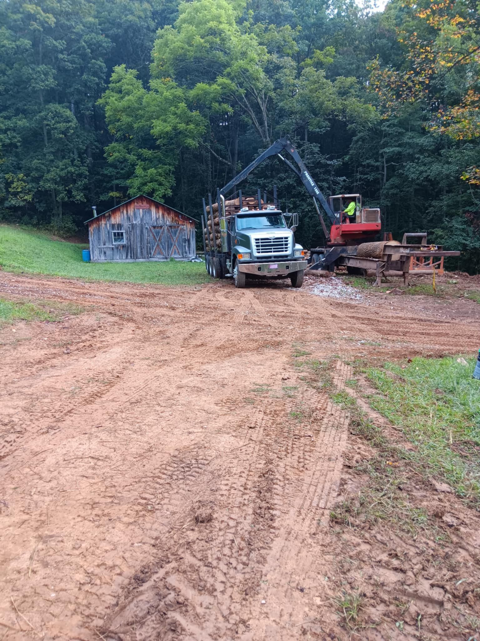Muddy dirt road with logging truck loading logs with a mechanical arm near a barn and forest.