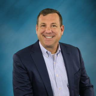 A man in a suit and striped shirt is smiling in front of a blue background.