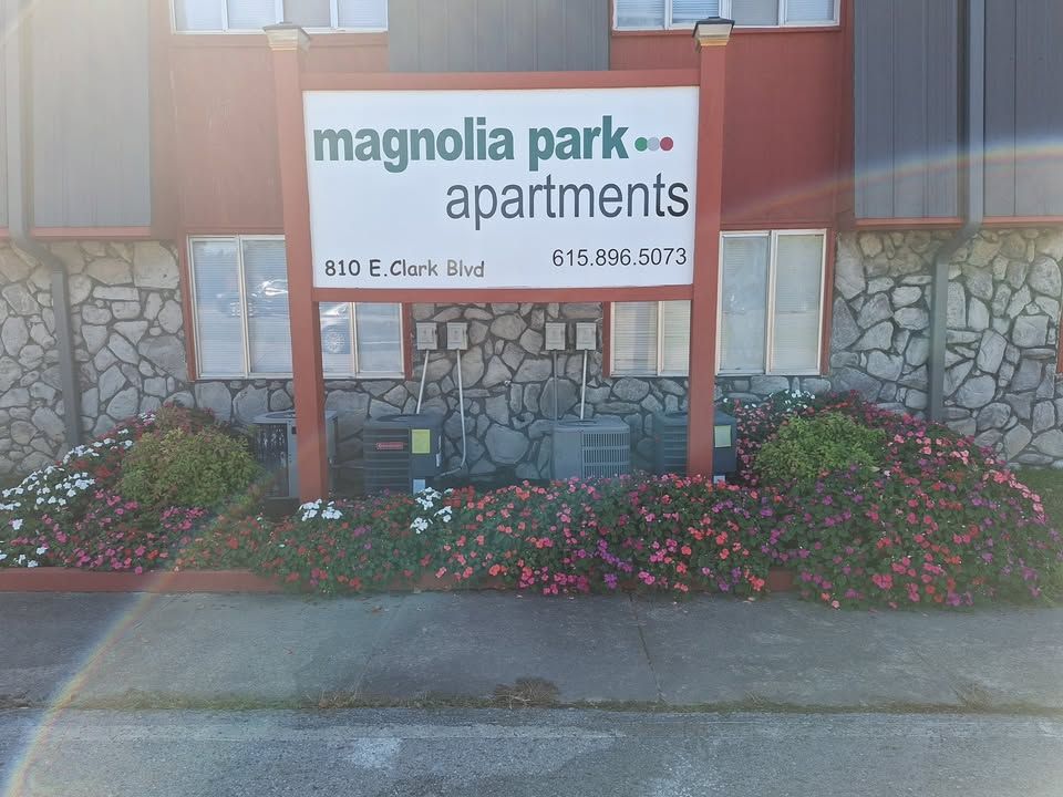 Sign for Magnolia Park Apartments at 810 E. Clark Blvd. with red and green text, surrounded by flowers.