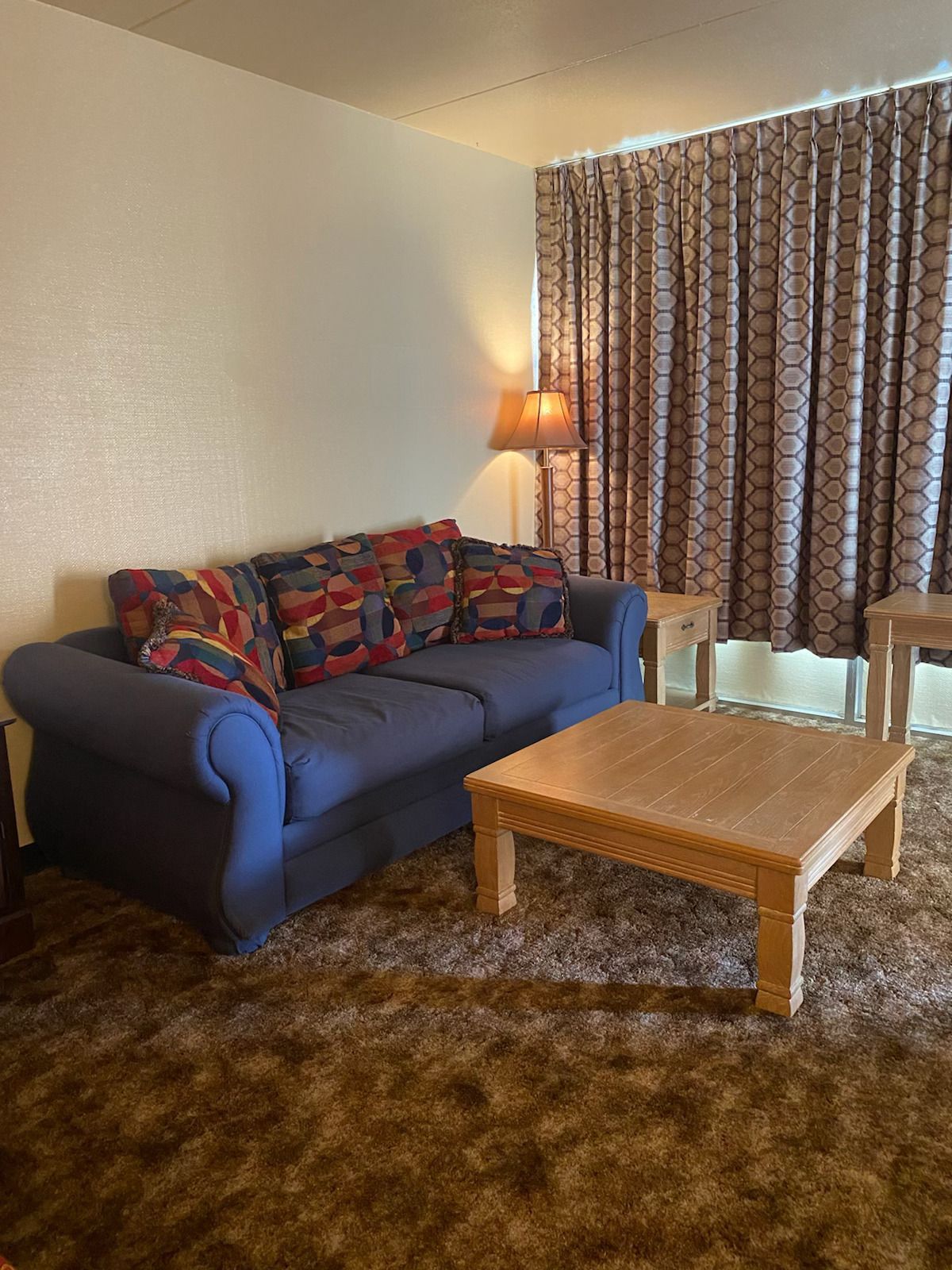 Blue couch with patterned pillows and a wooden coffee table in a room with patterned curtains and brown carpet.