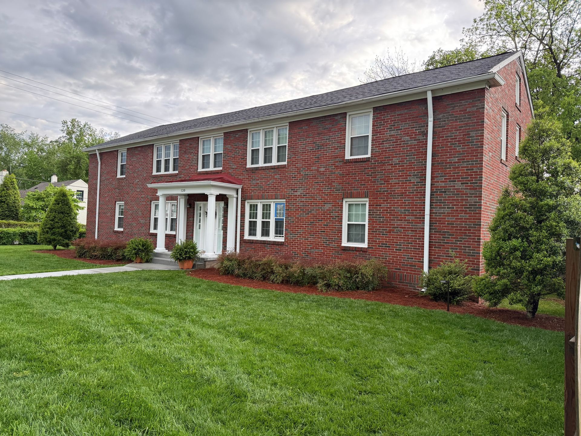 Two-story red brick apartment building with white columns and trim, green lawn, cloudy sky.