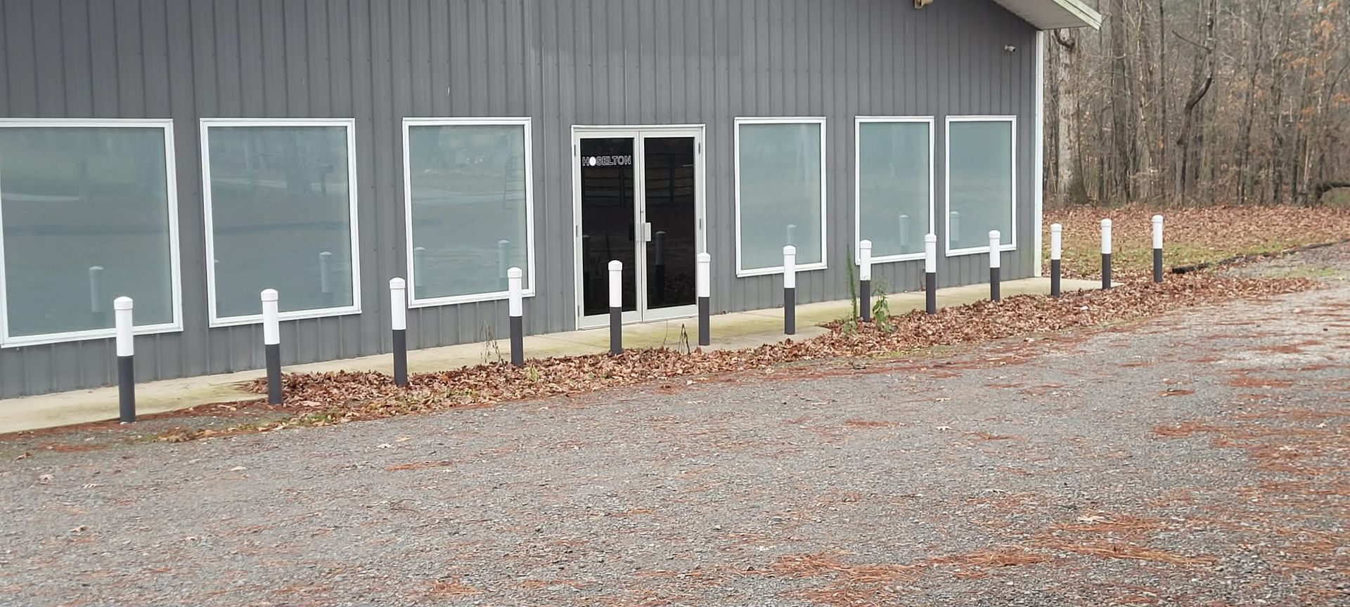 Gray building with windows and a gravel driveway. Black and white poles line the front.