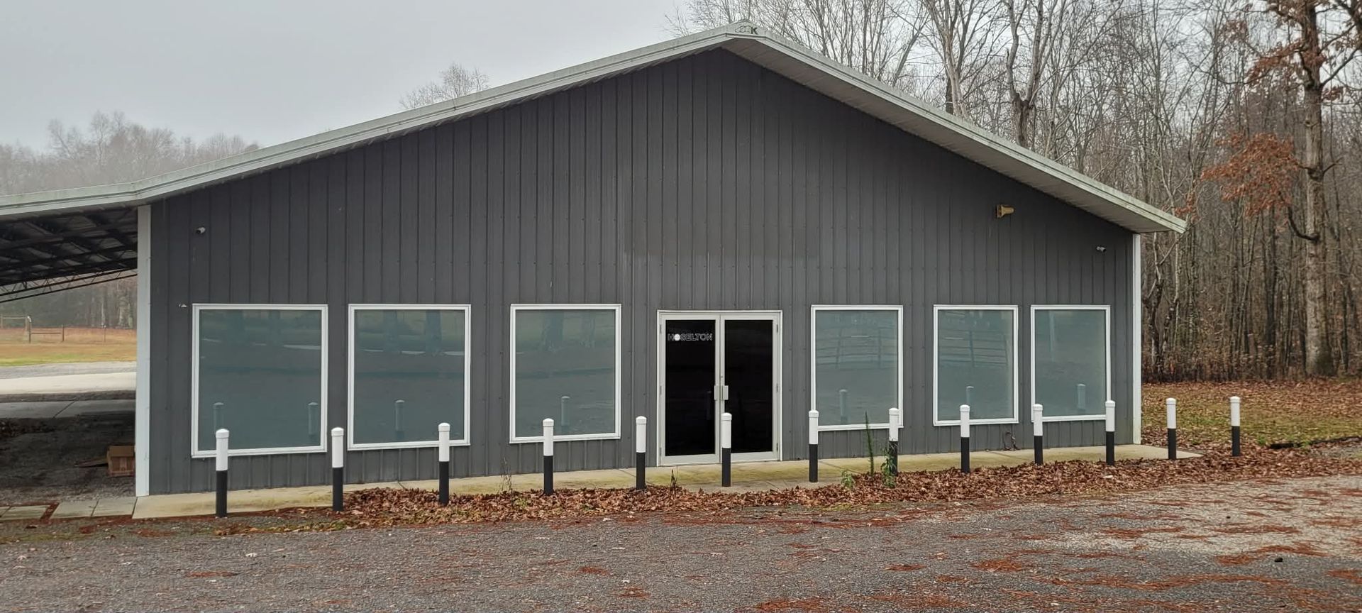 Gray building with large windows and a glass door, surrounded by trees and a gravel area.