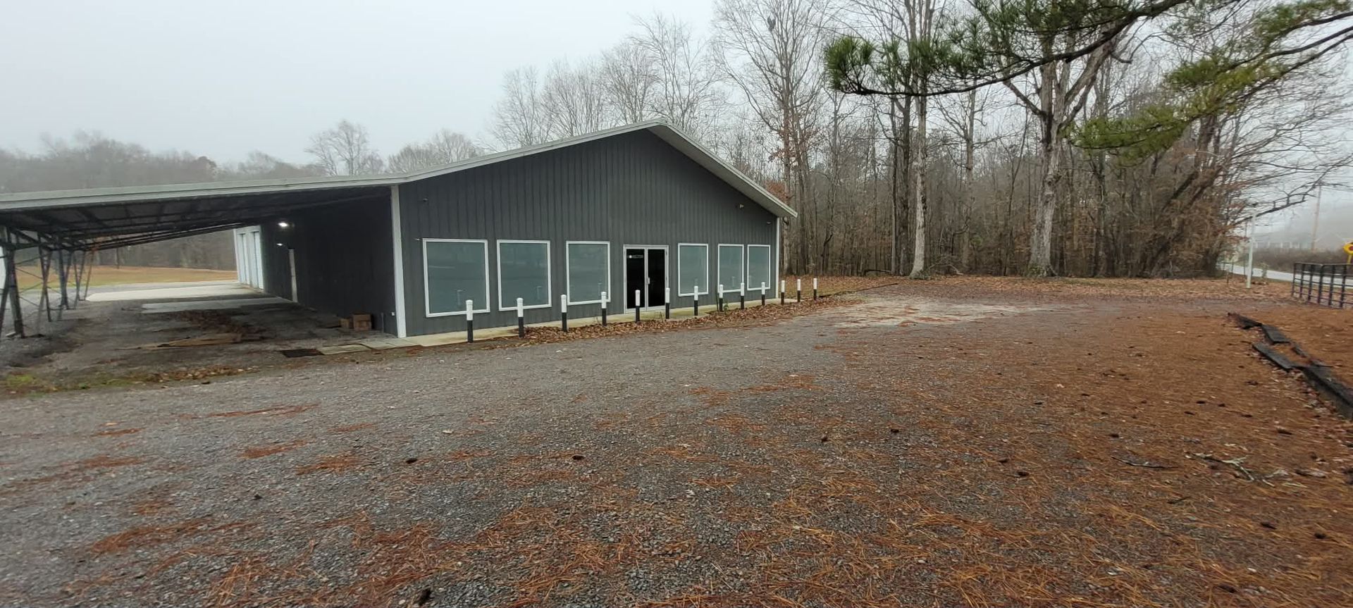 A dark gray building with several windows and a covered carport, set in a gravel area with bare trees in the background.