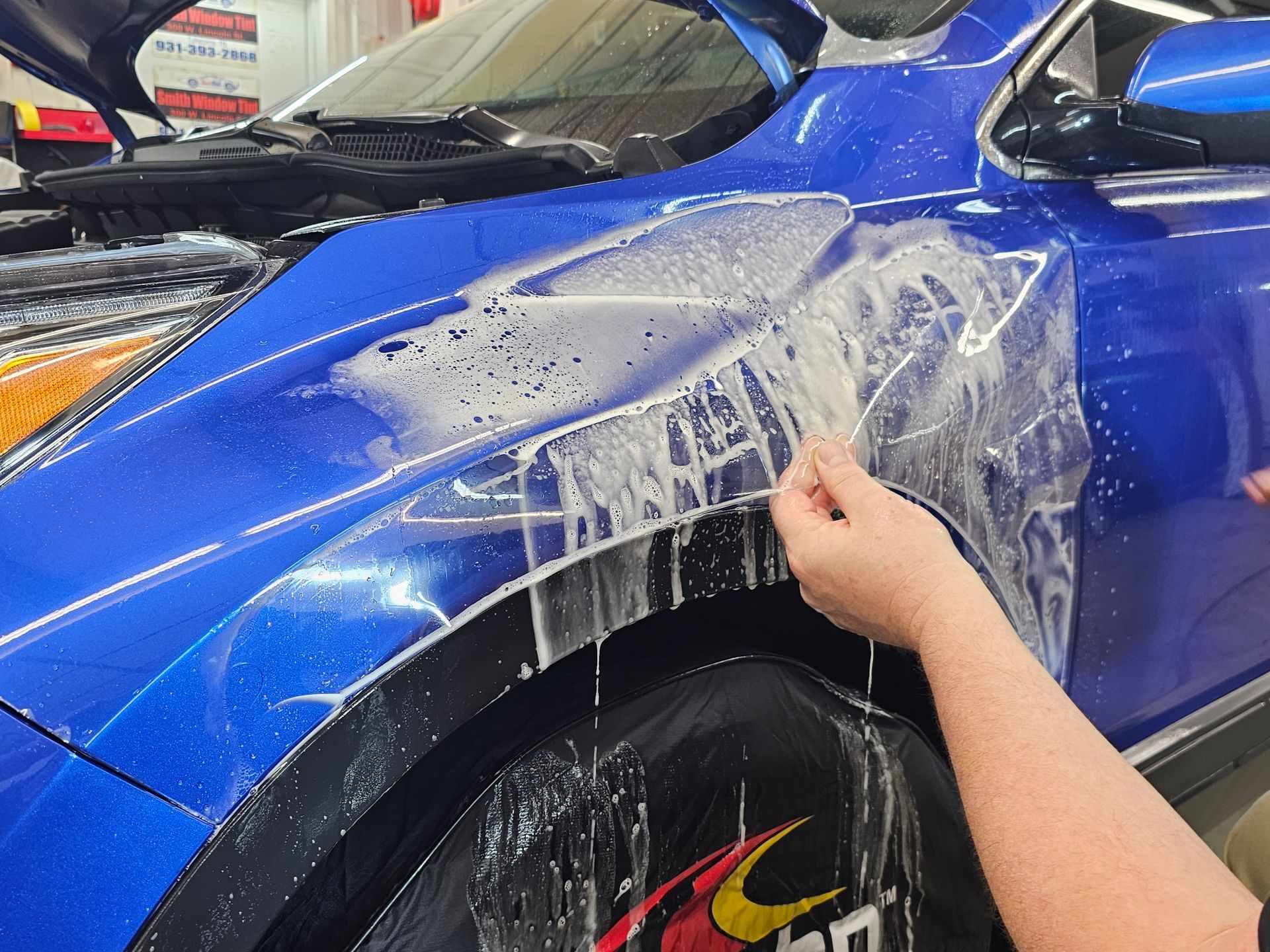 A person is applying a protective film to the fender of a blue car.