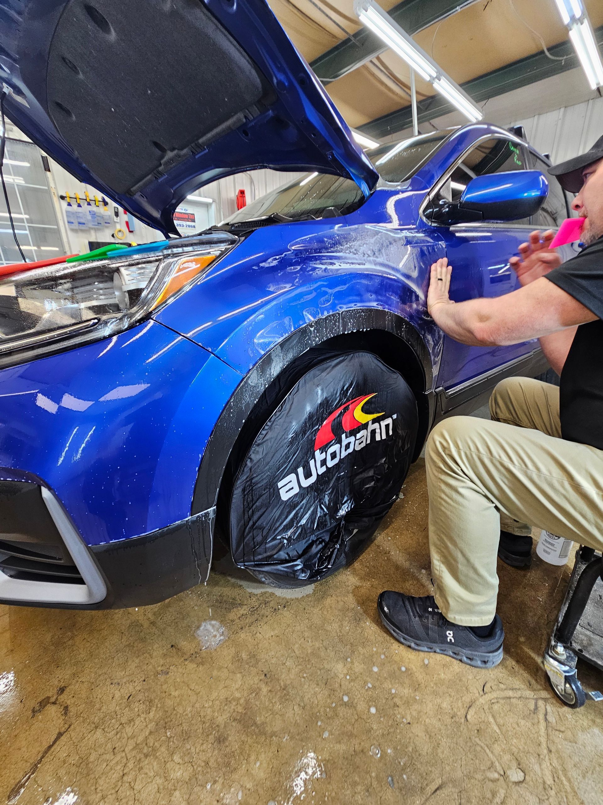 A man is kneeling down next to a blue car with the hood up.