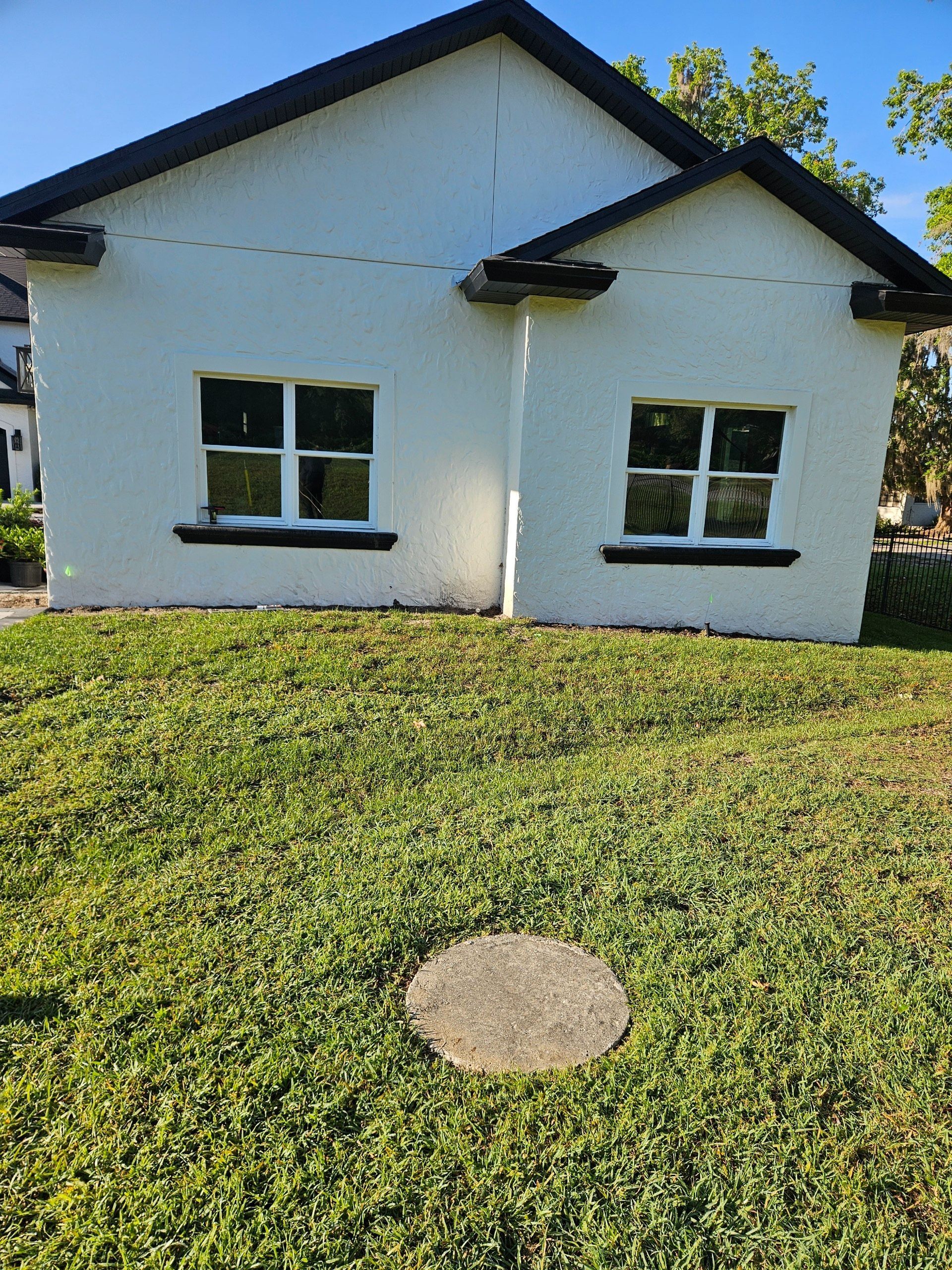 White building with two windows and a black roof, set on a grassy lawn with a circular stone.