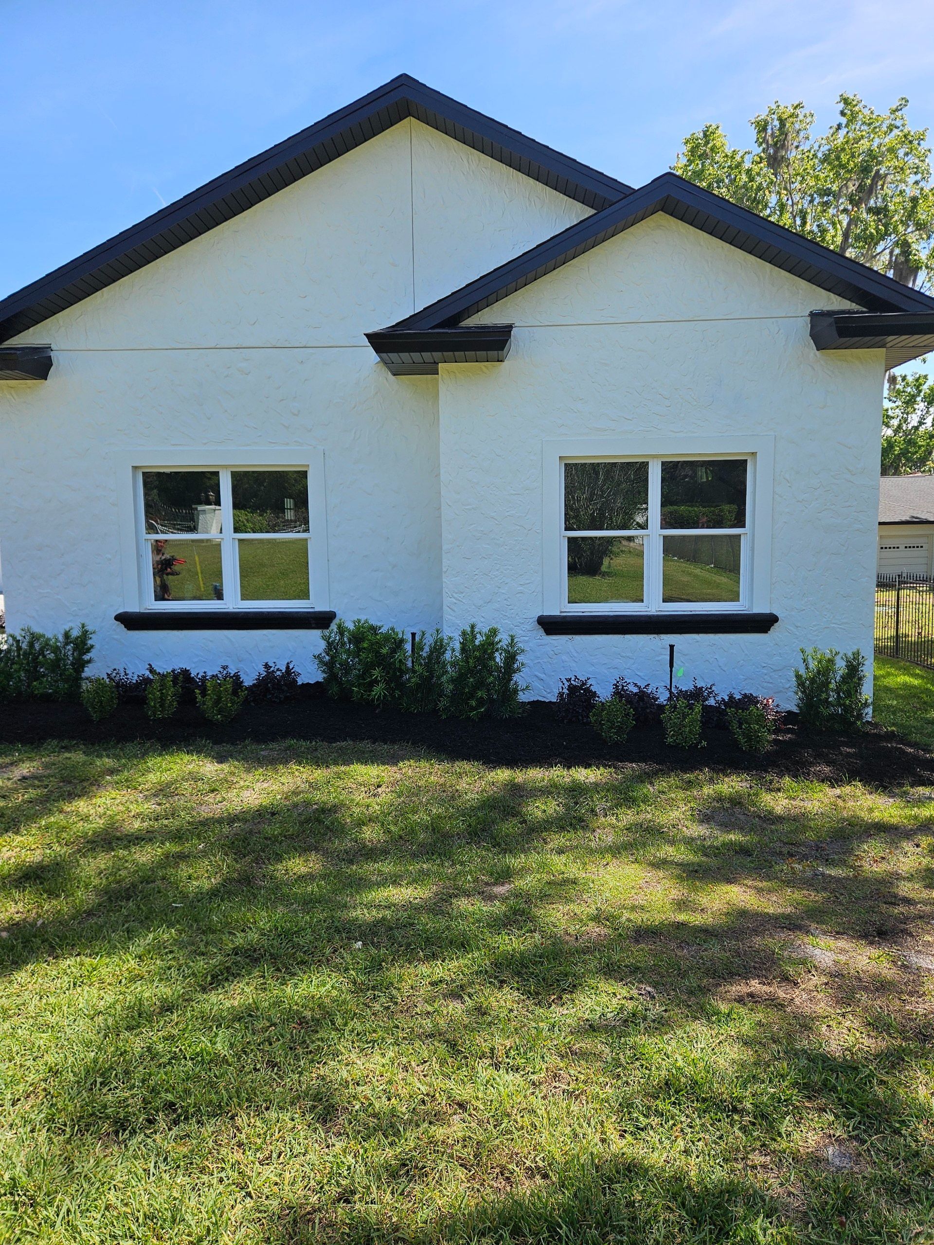 White house with black trim, two windows, and green landscaping. Blue sky.