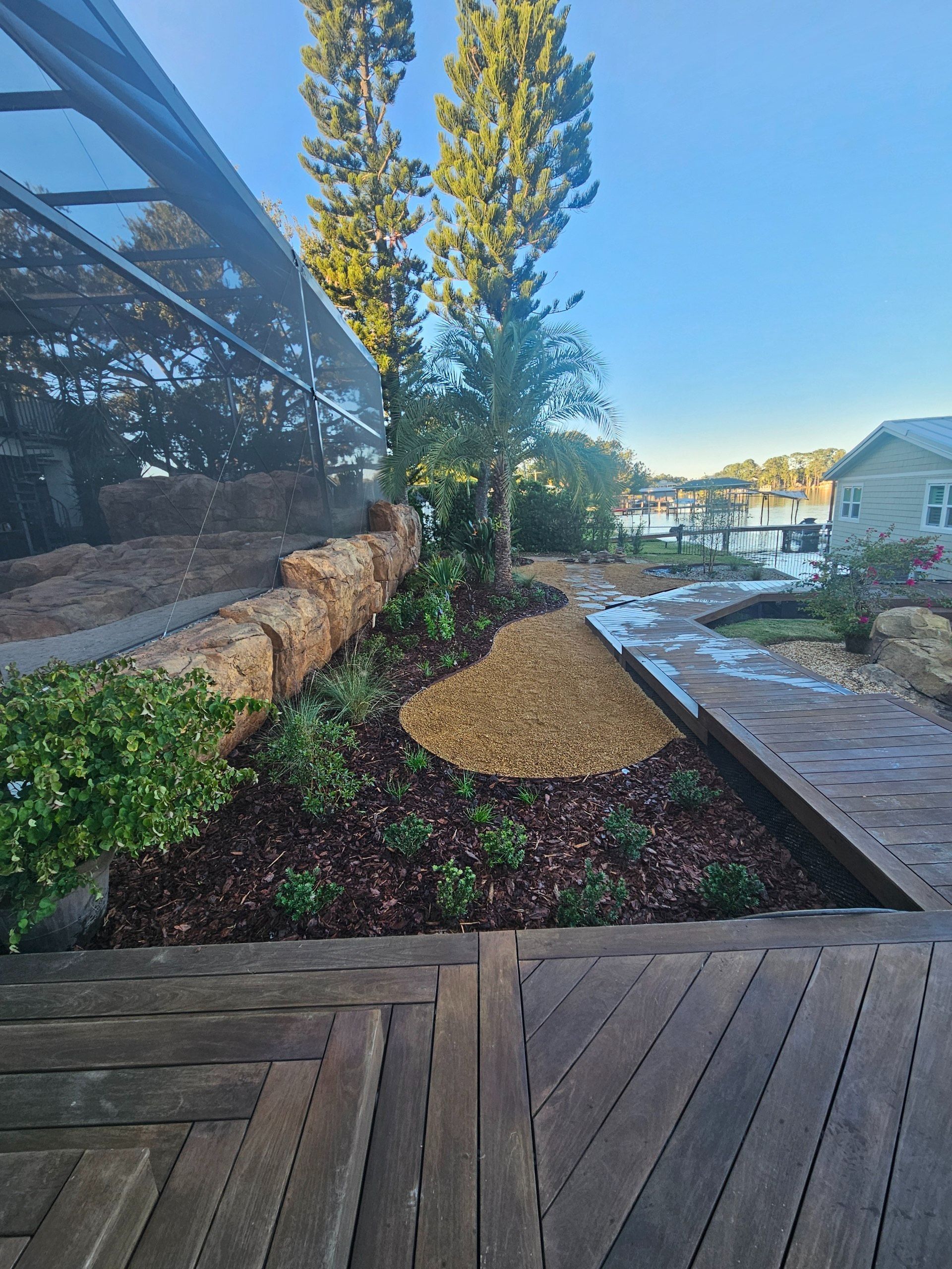 Landscaped garden with mulch, rocks, and a wooden deck. Trees and water in the background.