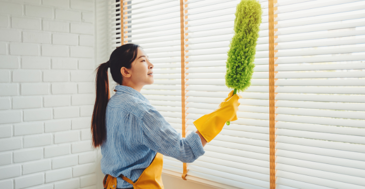 A woman is cleaning blinds with a duster.