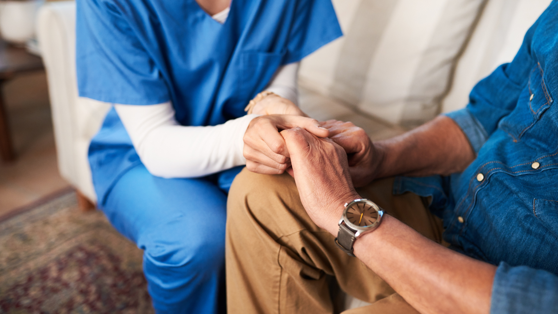 A nurse is holding the hand of an elderly man while sitting on a couch.