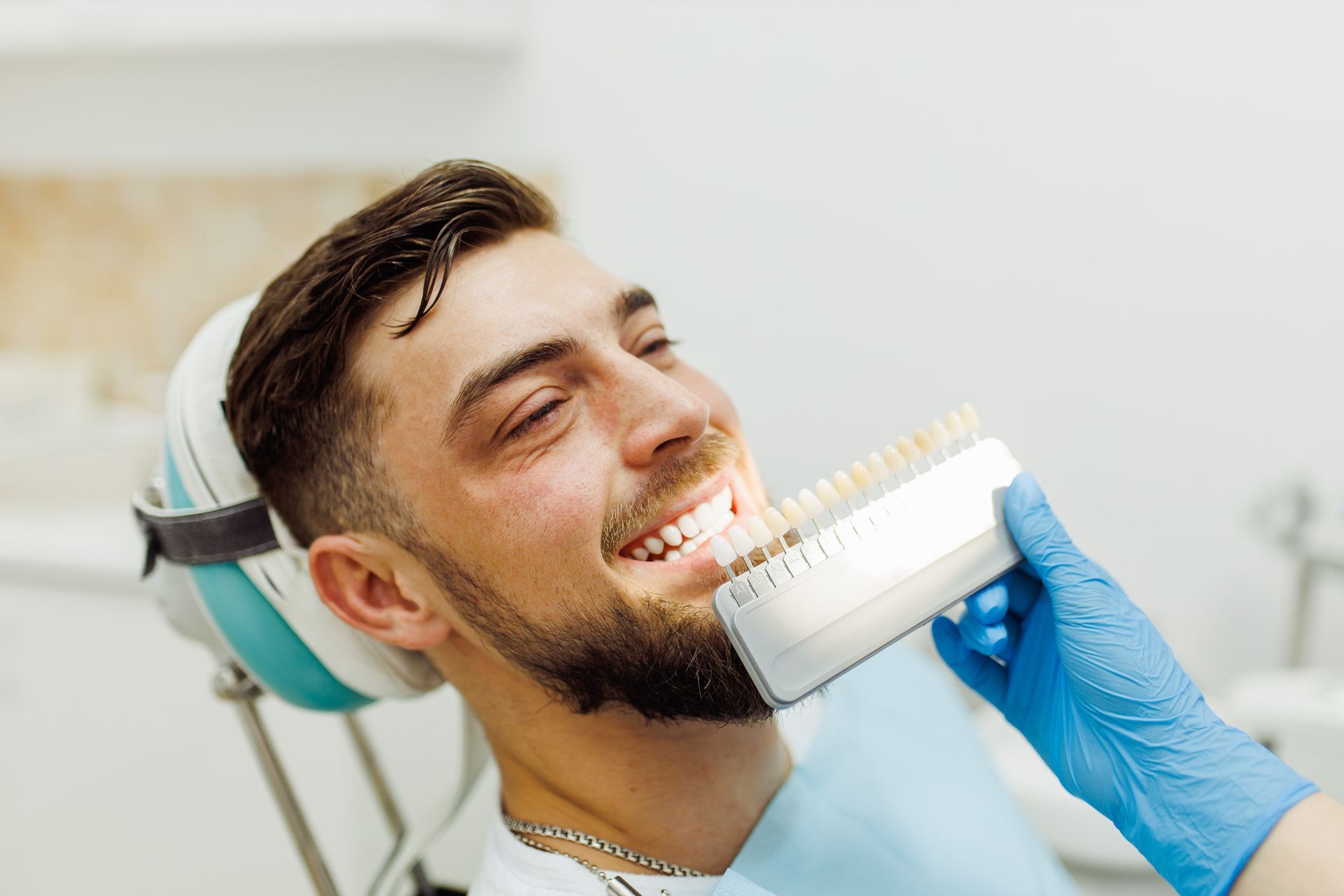 Young man sits in a dentist chair as a dentist holds a tooth color chart to his smile. Young man sits in a dentist chair as a dentist holds a tooth color chart to his smile.>