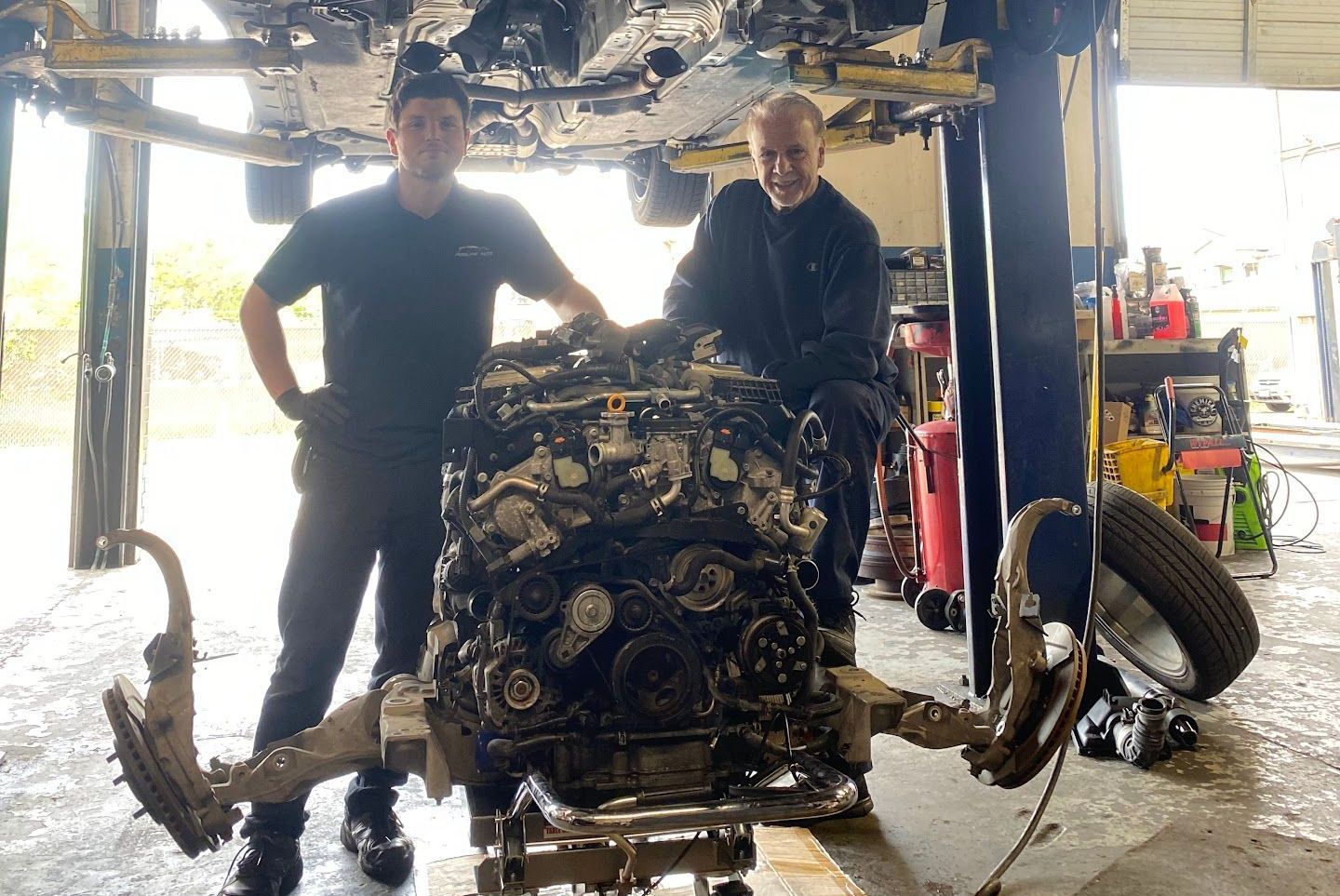 Two mechanics pose with a car engine removed from a vehicle in a repair shop. | Proline Auto