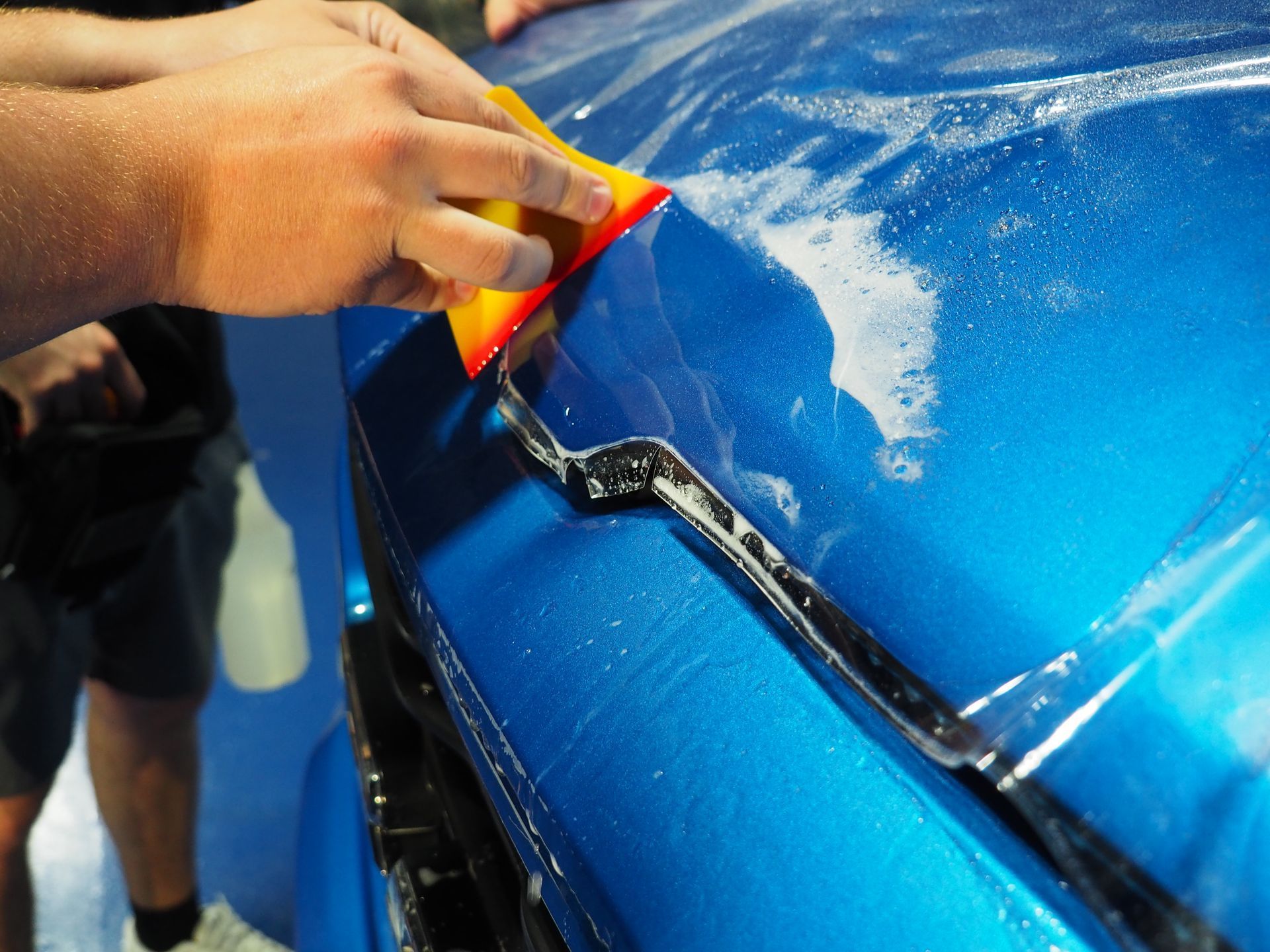 Person applies clear protective film to a blue car hood with a squeegee.